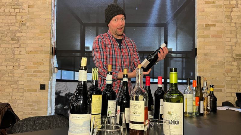 A man holding a bottle of wine behind a table of wine.